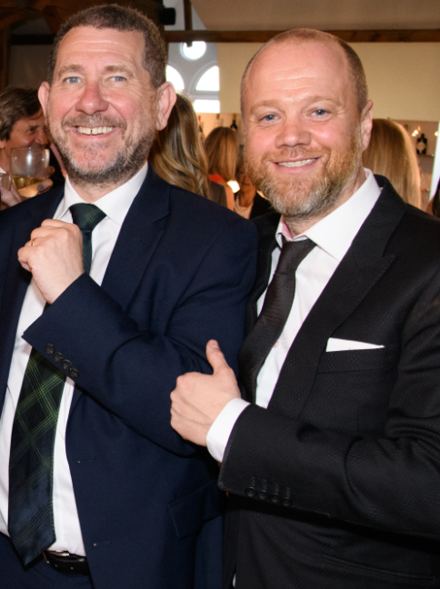 Two men in suits, standing and smiling warmly at camera. One has their thumbs up.