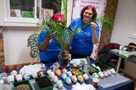 Image of two woman standing behind a plant and grinning. The plant is on a table covered in gardening equipment, to demonstrate a mental health gardening project run by one of the women.