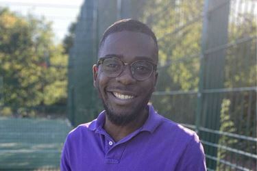 A young Black man wearing glasses and smiling, wearing a purple t-shirt and standing outdoors on a sport pitch on a sunny day