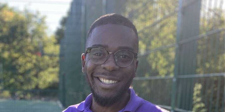 A young Black man wearing glasses and smiling, wearing a purple t-shirt and standing outdoors on a sport pitch on a sunny day