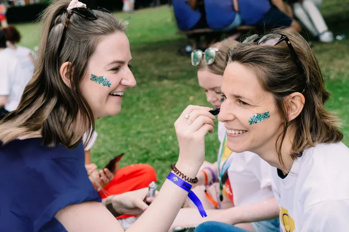 Two woman are sat smiling on the grass. One of the woman is doing the other's face glitter.