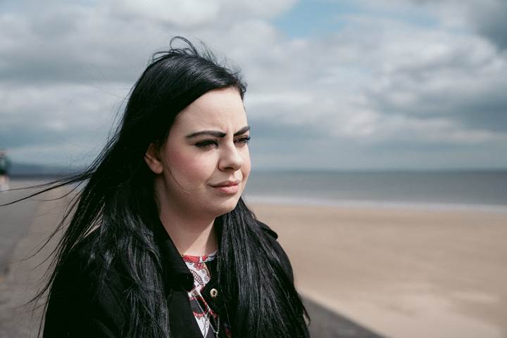 White woman with black hair looking to the side, with the beach in the background.
