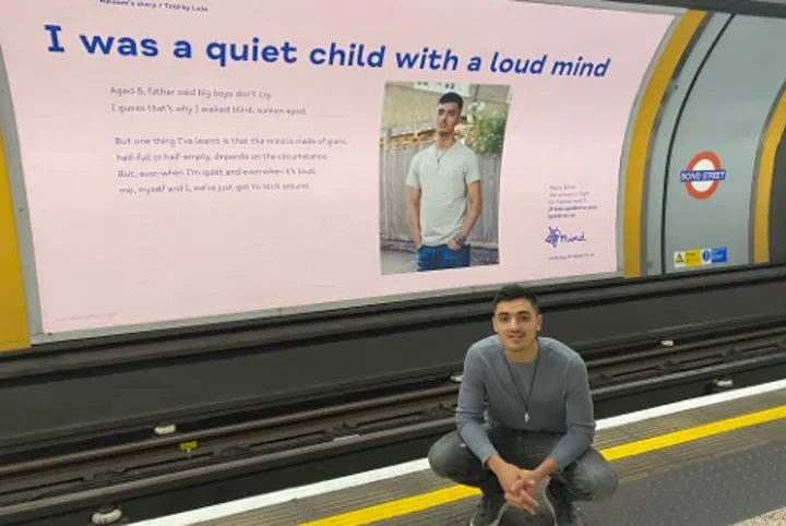 Media volunteer Haleem crouching outside a Mind billboard with his picture on a tube platform
