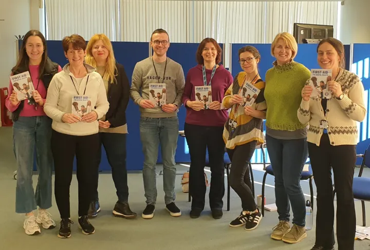 A group of people stand in a row, all smiling to the camera, holding up 'First Aid' leaflets.