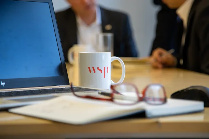 A desk with a laptop, a pair of red reading glasses, a WSP mug, and some paperwork on it. We can see sat at the table in the background.