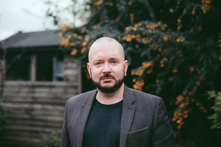 White bald man with dark brown beard, looking at the camera. Garden in the background.