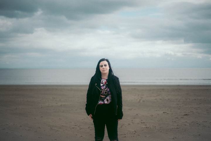 White woman with black hair standing on the beach, looking directly at the camera.