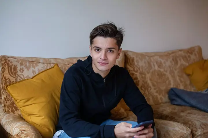 Young person with short brown hair looks at the camera smiling while using a phone. They are seated on a light brown sofa with yellow cushions.