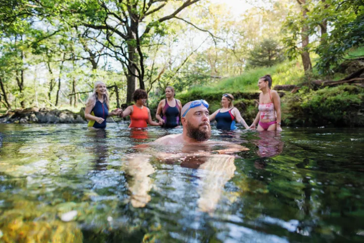 A group of friends are swimming together in a small lake. Behind them you can see tall trees, and light green foliage, with the sun shining across the backdrop.